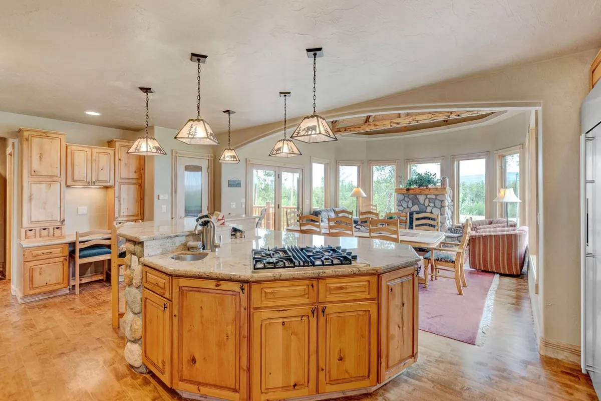 A bright, welcoming kitchen scene showcasing a central kitchen island with bar seating, stainless steel appliances, large windows letting in ample natural light, and a cozy dining area adjacent to the kitchen. The stone and wood finishes evoke warmth and sophistication, and the island offers a space for both cooking and socializing.