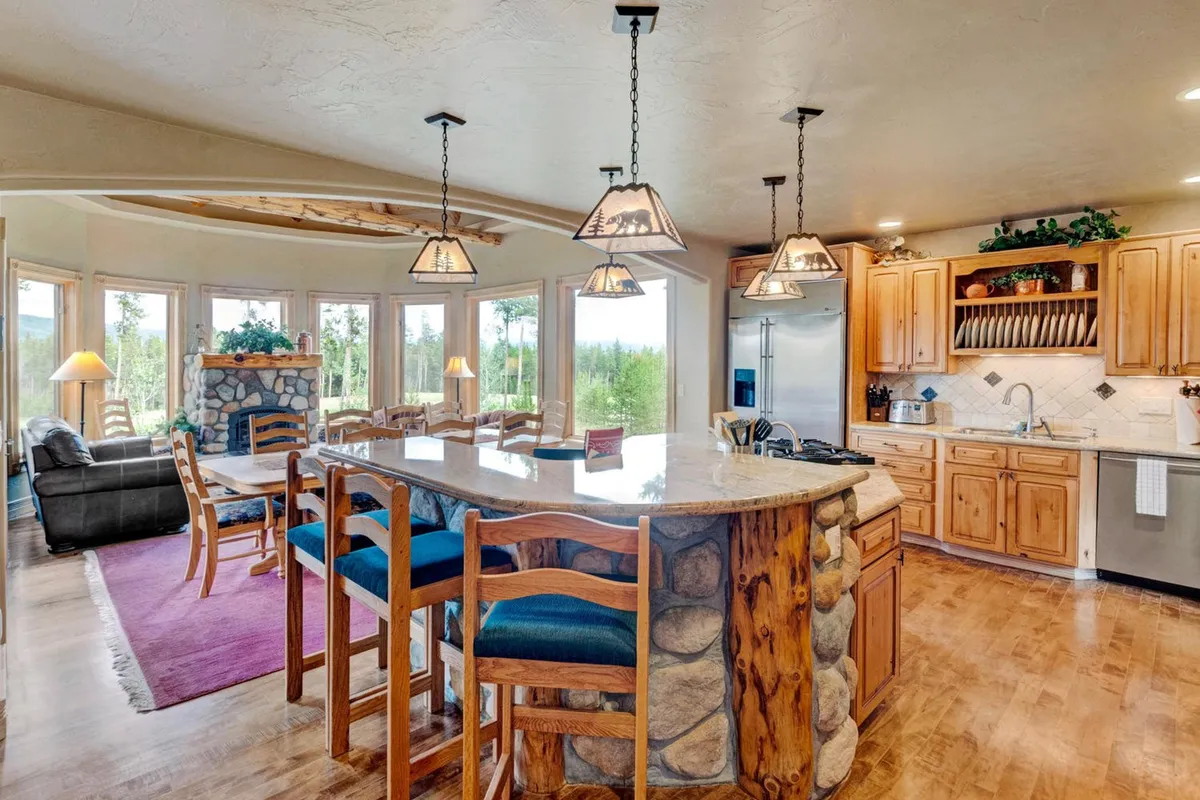 A panoramic view of the open kitchen and dining area, featuring a large stone fireplace, expansive windows with mountain views, an island with bar seating, and a spacious dining table. The space is filled with natural light and designed for both small gatherings and large celebrations.
