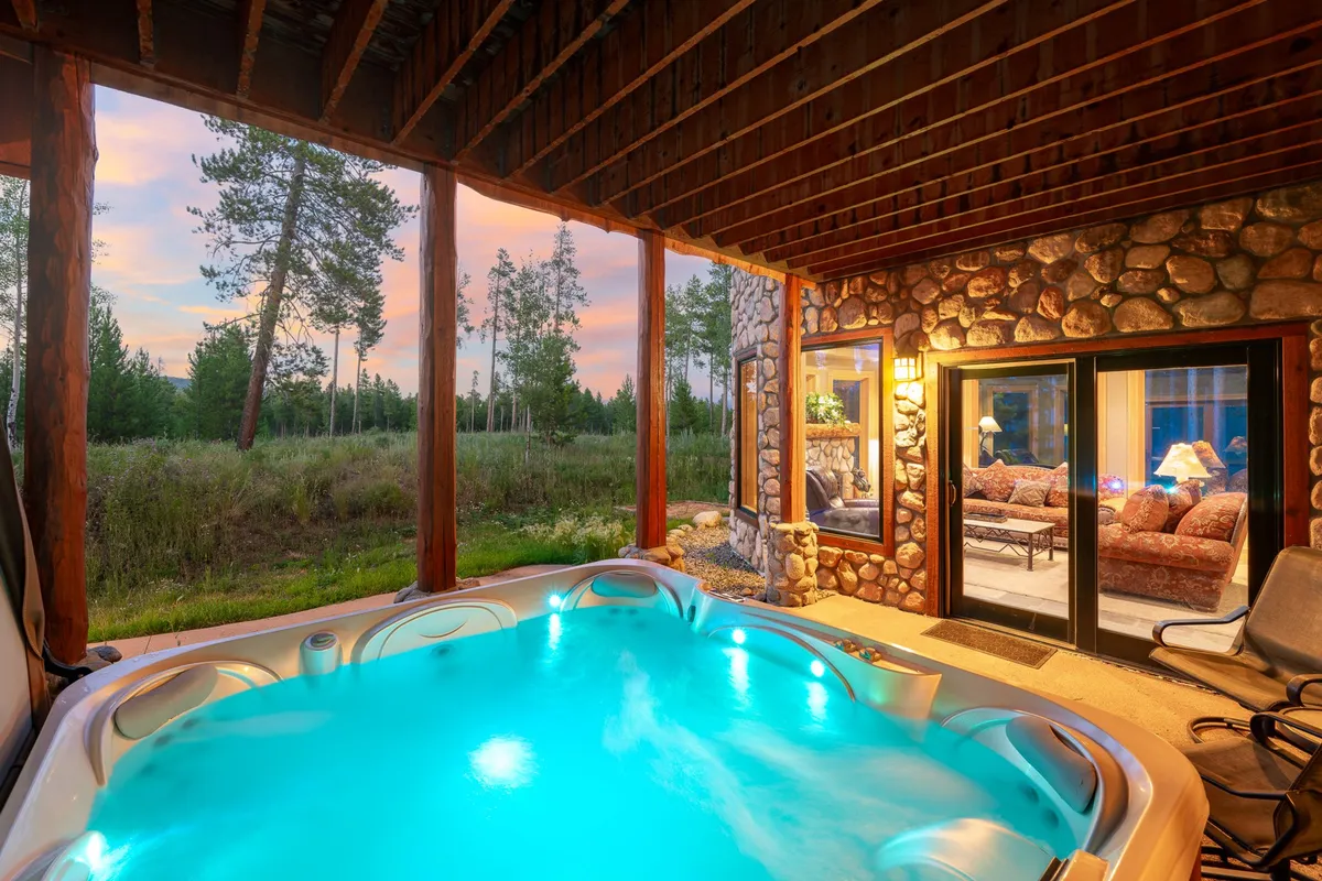 An outdoor hot tub illuminated with glowing lights, set against a backdrop of forest and a brilliant sunset. Comfortable seating is visible through a nearby glass door, emphasizing the connection between indoor and outdoor relaxation.