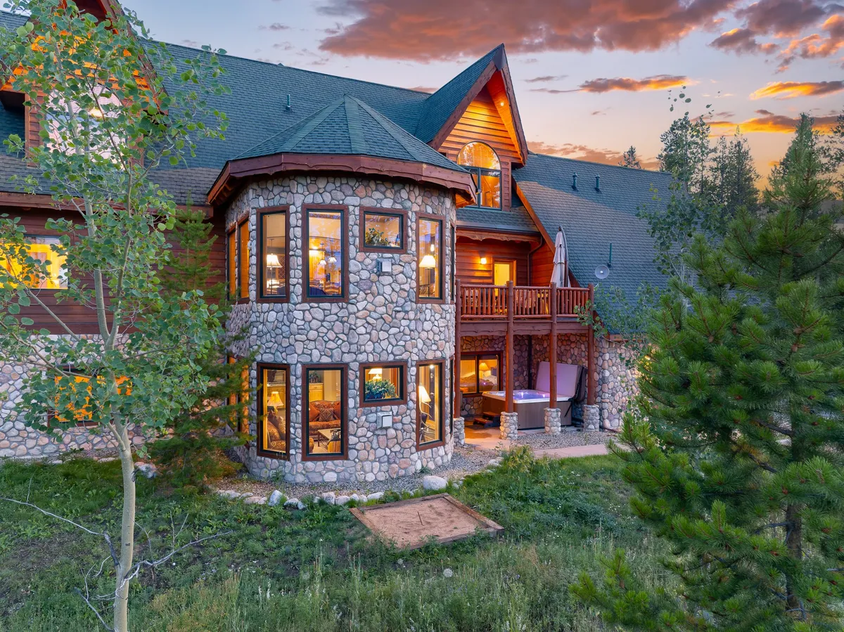 A striking view of the chalet’s exterior, featuring a distinctive stone turret, wooden balcony, expansive windows, and lush surrounding trees. The scene suggests inviting outdoor spaces perfect for family gatherings and relaxation in nature.