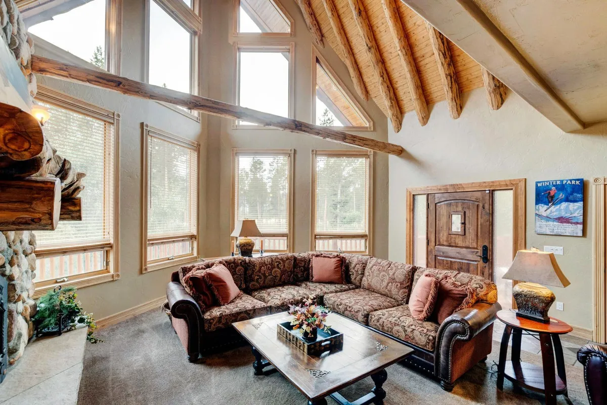 A welcoming living area with a large sectional sofa, a rugged stone fireplace, floor-to-ceiling windows, and exposed wooden beams. The scene is set for family or group gatherings, with abundant natural light and scenic views.