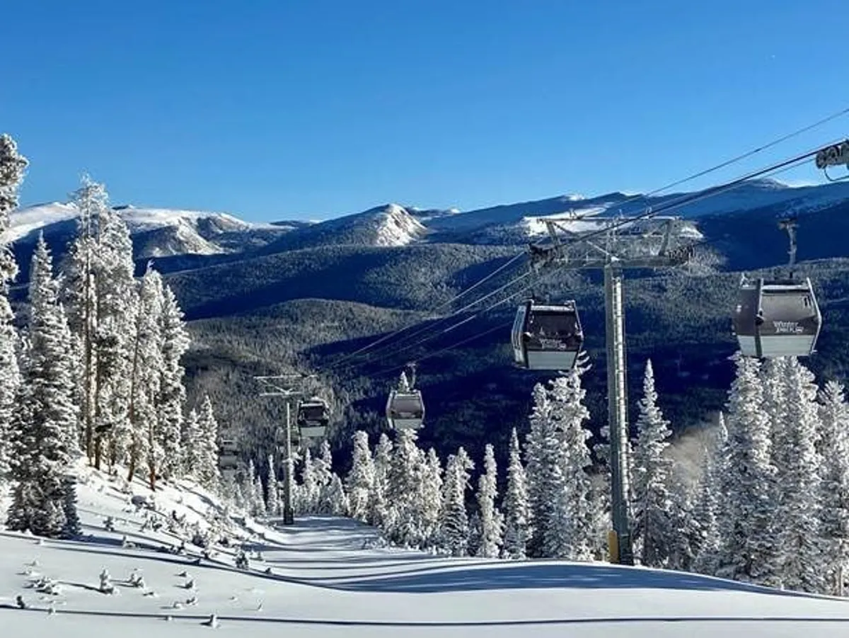 A picturesque exterior view of a ski lift ascending over snow-covered trees and mountain slopes beneath a clear blue sky. The scene exudes adventure and the beauty of the Rockies in winter.