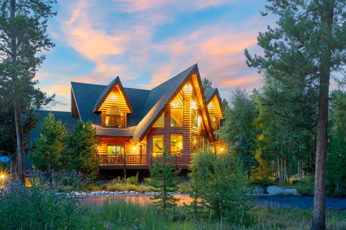 The image showcases a stunning mountain home with an intricate roofline, warmly lit windows that create a welcoming glow, lush greenery surrounding the property, and inviting outdoor spaces perfect for family gatherings or relaxing evenings watching the sunset.