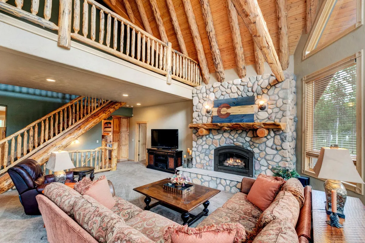 A soaring, light-filled great room featuring a two-story ceiling, plush sofas, a cozy stone fireplace, and expansive windows offering mountain views. The arrangement encourages group relaxation and conversation, with a staircase and loft area adding architectural interest.