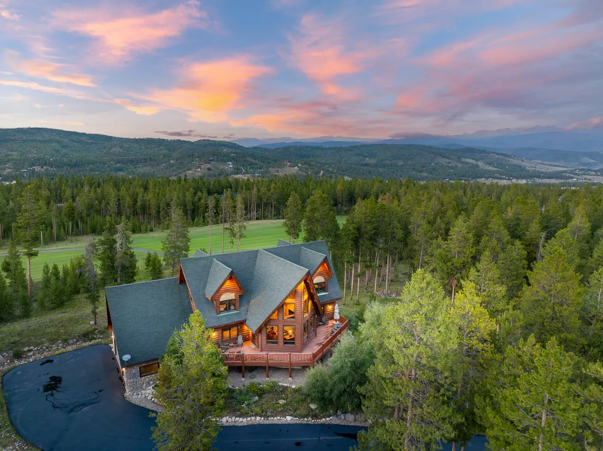 This exterior image features a picturesque log cabin house set against a vibrant sunset sky, surrounded by expansive green landscape and dense forest. The spacious deck and the house's inviting architecture are highlighted, with plenty of room for outdoor gatherings or quiet stargazing, all under the vast Colorado sky mentioned in the section.