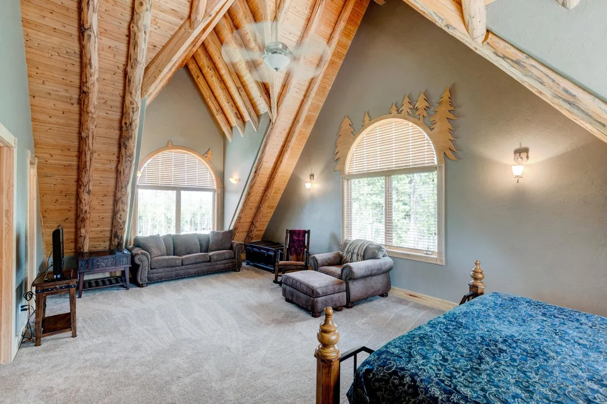 A spacious bedroom with an ornate bed, vaulted wooden ceiling, large windows framing mountain views, and comfortable seating. The serene ambiance is enhanced by natural light, creating a tranquil, upscale sanctuary.