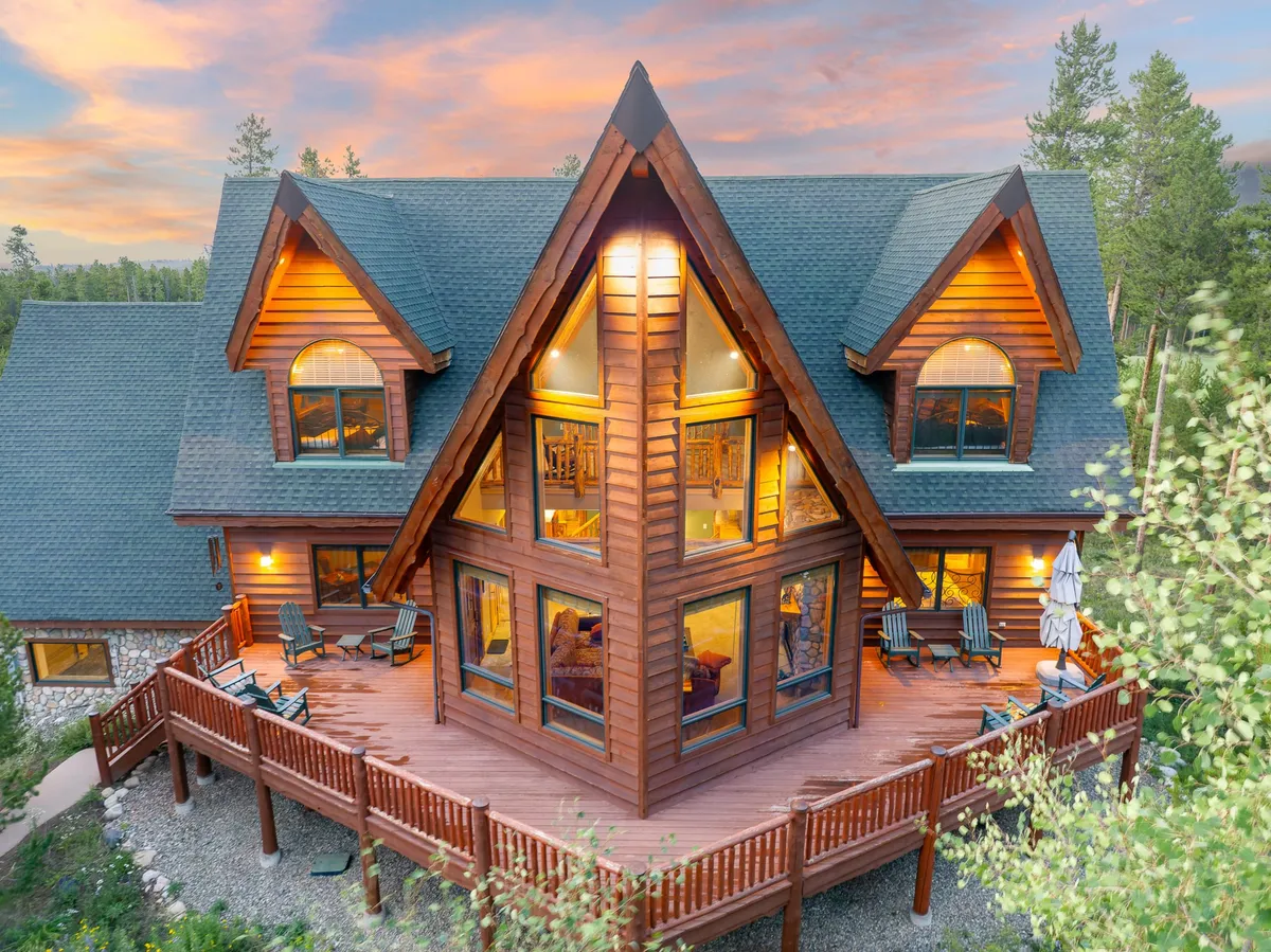 The chalet’s striking A-frame exterior, featuring impressive windows and a generous wraparound deck nestled among trees. The deck is set up for outdoor dining or group relaxation, seamlessly connecting inside and outside living.