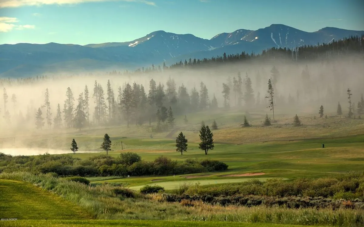 A tranquil view of the golf course stretching out with early morning fog lifting over the greens, framed by trees and rugged mountain peaks. The image suggests opportunities not only for golf but also for hiking, nature walks, and wildlife viewing.